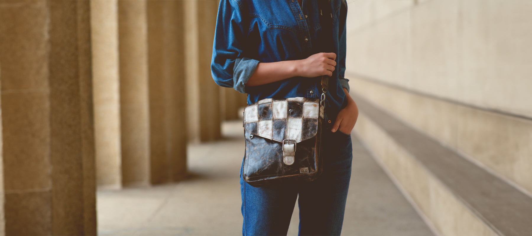 A woman in denim shirt and pants holding a blue and gray tie-dyed wallet with a rustic metal backdrop.