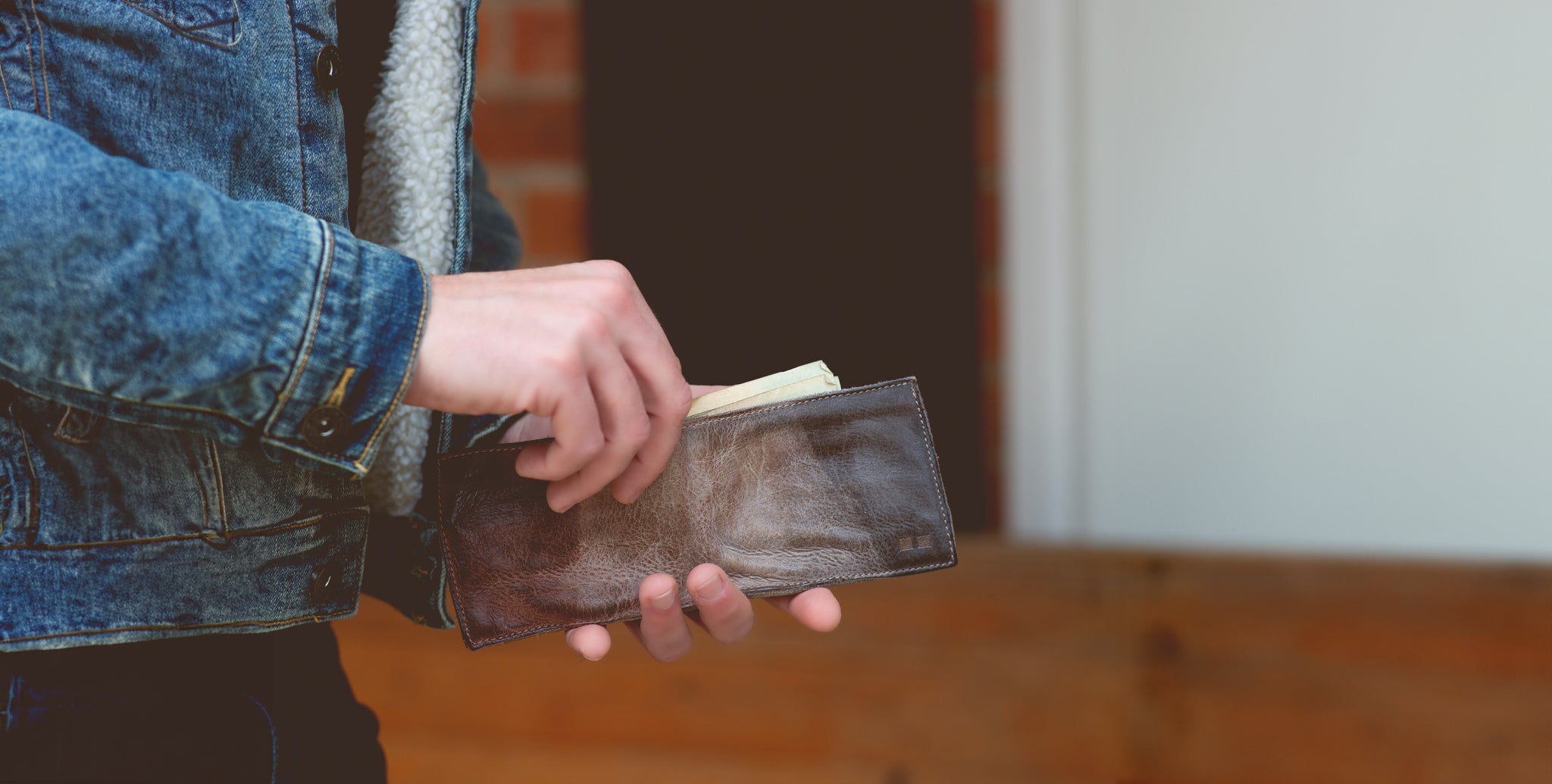 A person in a denim jacket taking cash out of a brown leather wallet.