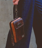 A person holds the Alelike, a small compact crossbody phone holder made of premium leather by Bed Stu, by a wrist strap, wearing dark blue jeans against a plain background.