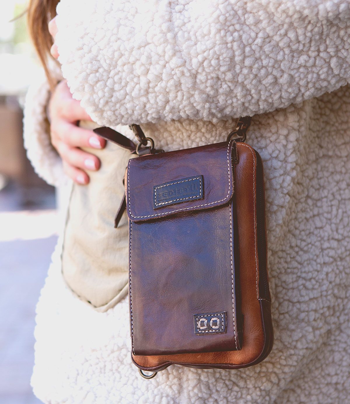 A person wearing a fuzzy jacket is holding the Alelike premium leather crossbody bag by Bed Stu, featuring a front flap and stitching details, showcasing its daily functionality as a compact phone holder.