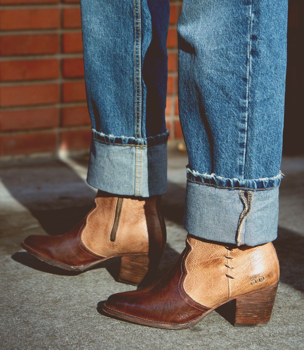 A person wearing rolled-up blue jeans and the Bed Stu Baila II brown leather ankle boots with chunky heels and whipstitch accents stands on a concrete surface against a brick wall backdrop, exuding cowgirl vibes.