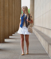 A woman stands on a stone path between columns, wearing a light brown shirt, blue vest, white skirt, and Bed Stu Ballet shoes designed for comfort and flexibility, holding a beige bag.
