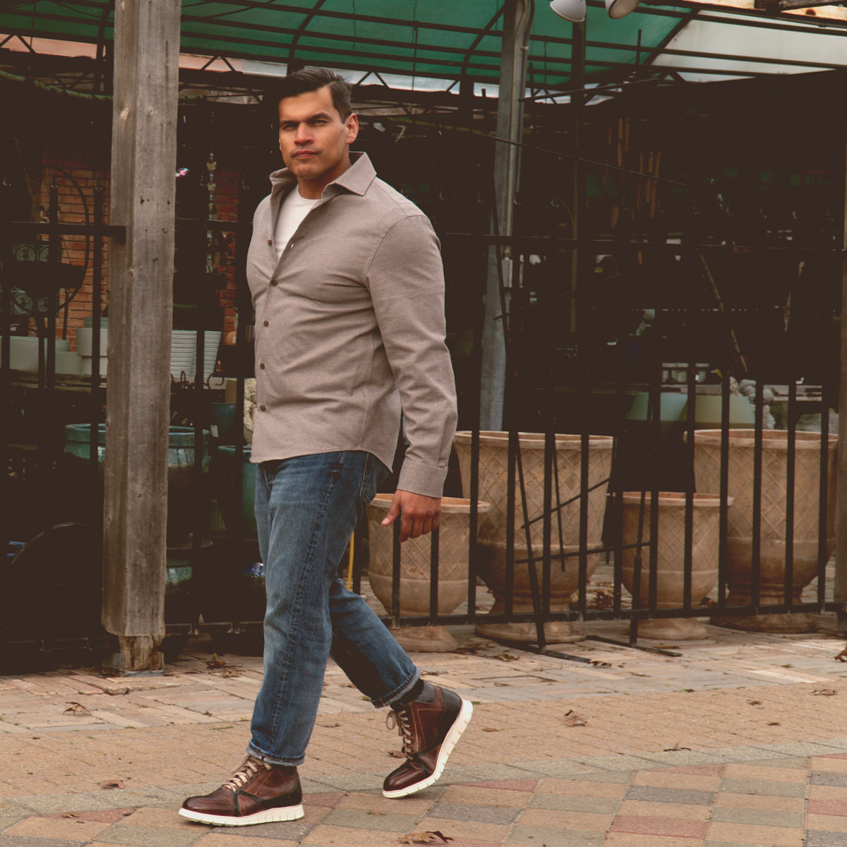 A man strolls on a paved pathway in a gray shirt and jeans, flaunting his Bed Stu Barton leather high-top sneakers. Potted plants and a wooden structure enhance the scene's charm while he relishes the comfort of breathable insoles with each step.