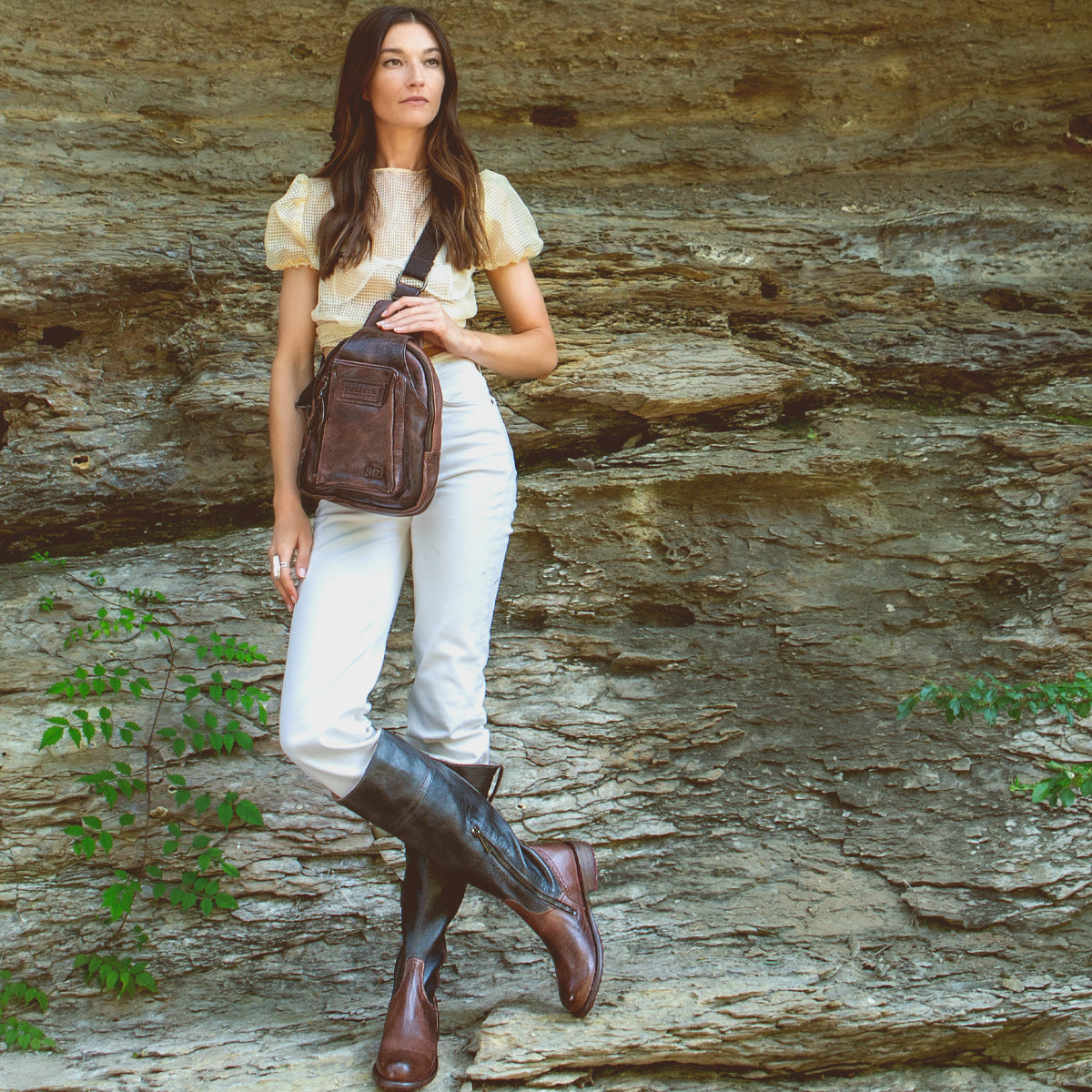 A woman is standing on a rock with a Beau sling backpack from Bed Stu, which features a durable strap holding the leather backpack.