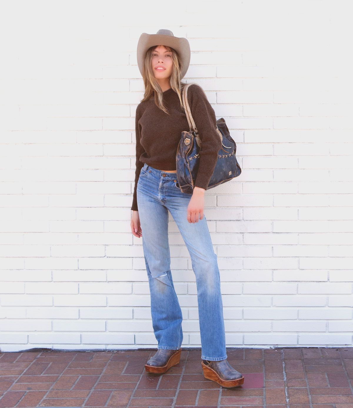 A woman with long blonde hair, wearing a black sweater, blue jeans, brown shoes, and a gray cowboy hat, stands against a white brick wall holding an artisanal Bruna denim bag by Bed Stu.