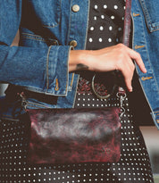 A person holds the Bed Stu Cadence, a maroon and black leather crossbody bag, in front of a black and white polka dot dress paired with a denim jacket.