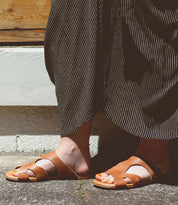 A person wearing Bed Stu's Configure brown leather sandals with a cushioned inner sole and a black and white striped skirt stands on a cement ground.