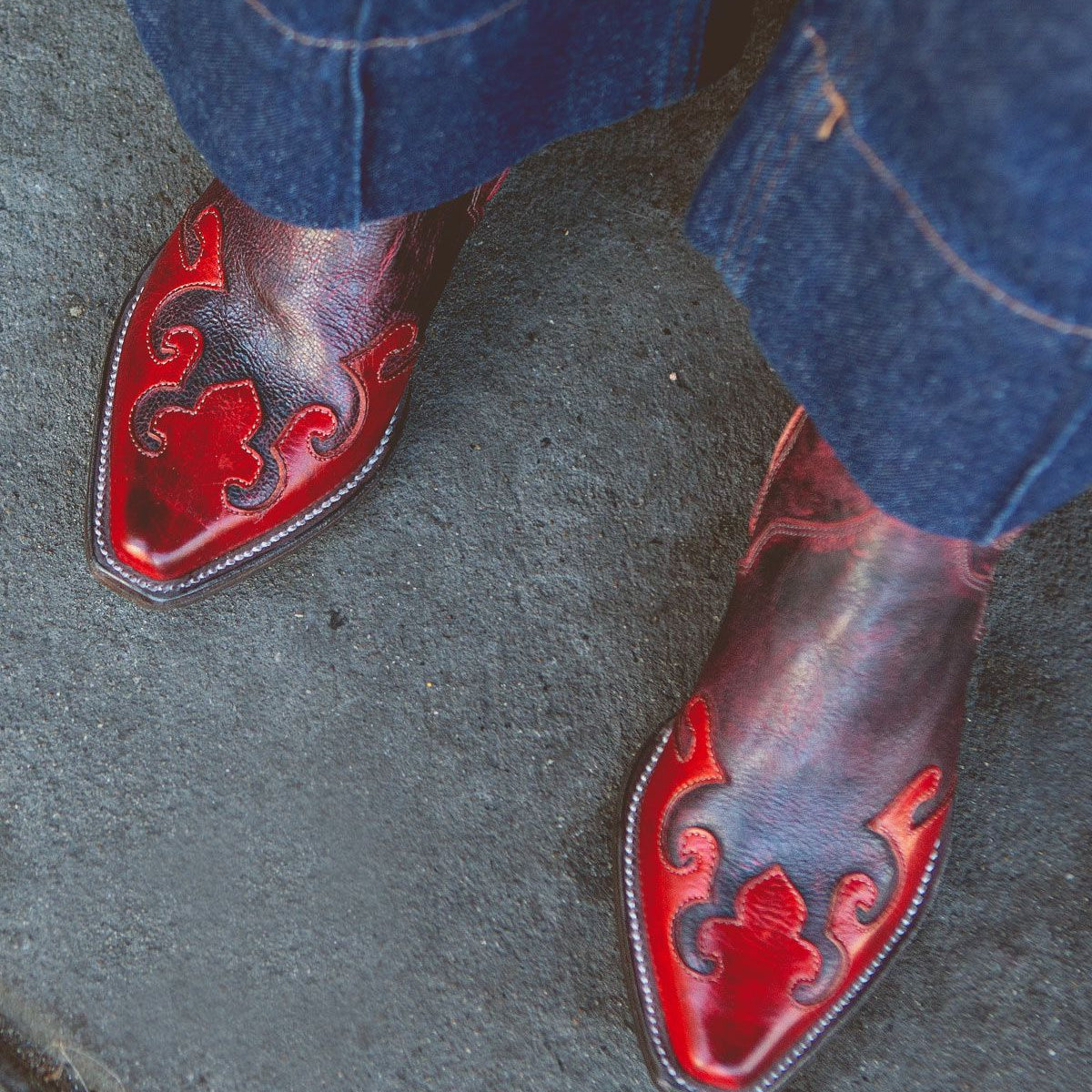 A pair of shiny red and black Deuce women's mid-calf cowboy boots by Bed Stu, featuring decorative stitching, worn with blue denim pants, showcasing a classic western style.