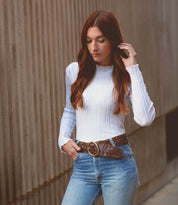 A person with long brown hair, exuding vintage allure, wears the Dreamweaver sweater by Bed Stu along with jeans while standing near a textured wall.