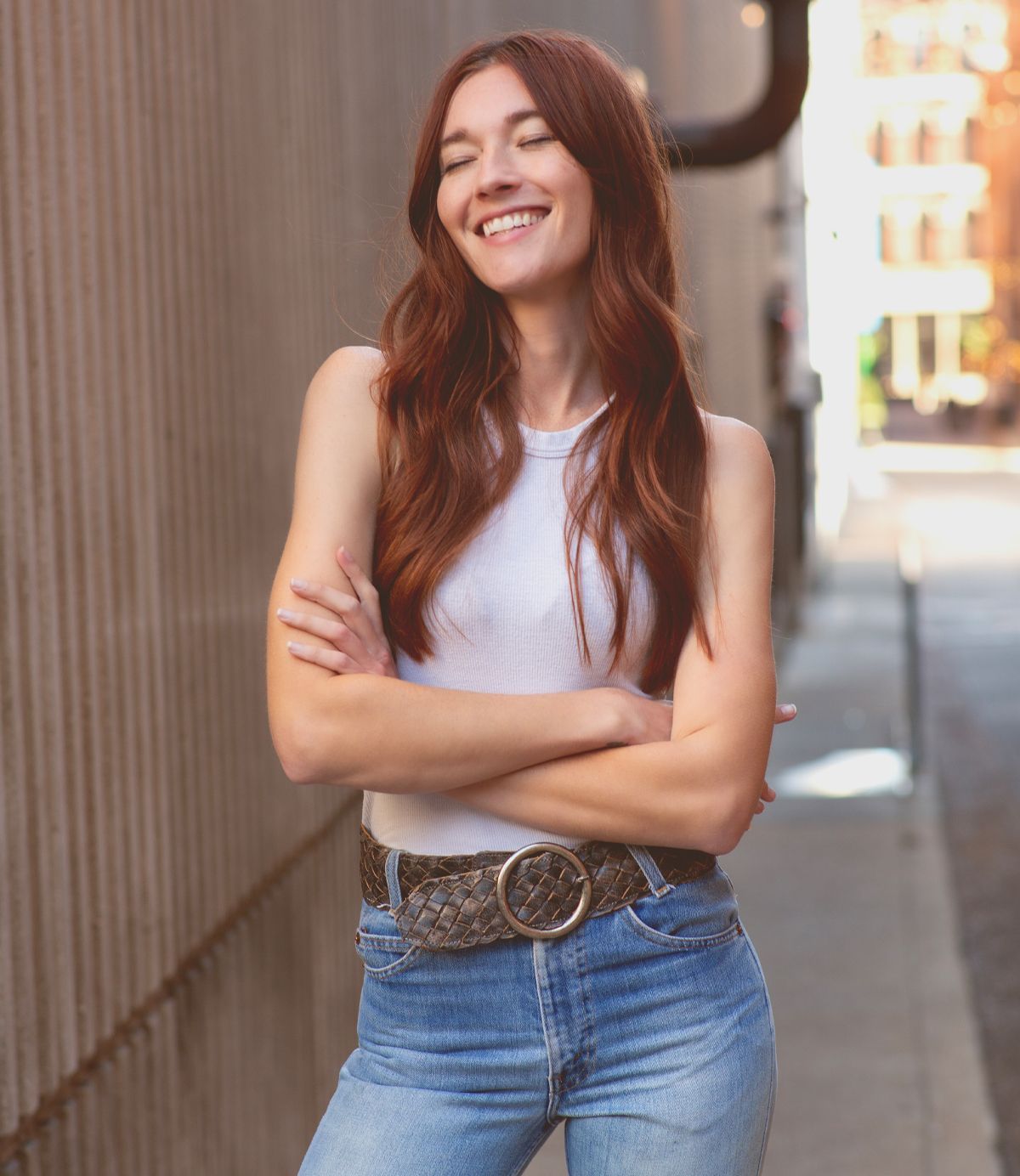 A woman with a vintage allure, long red hair cascading down her shoulders, stands in an alley with arms crossed, wearing a white tank top and jeans. She smiles subtly, her look accentuated by the Bed Stu Dreamweaver brass-tone buckle that adds a hint of timeless charm to the scene.