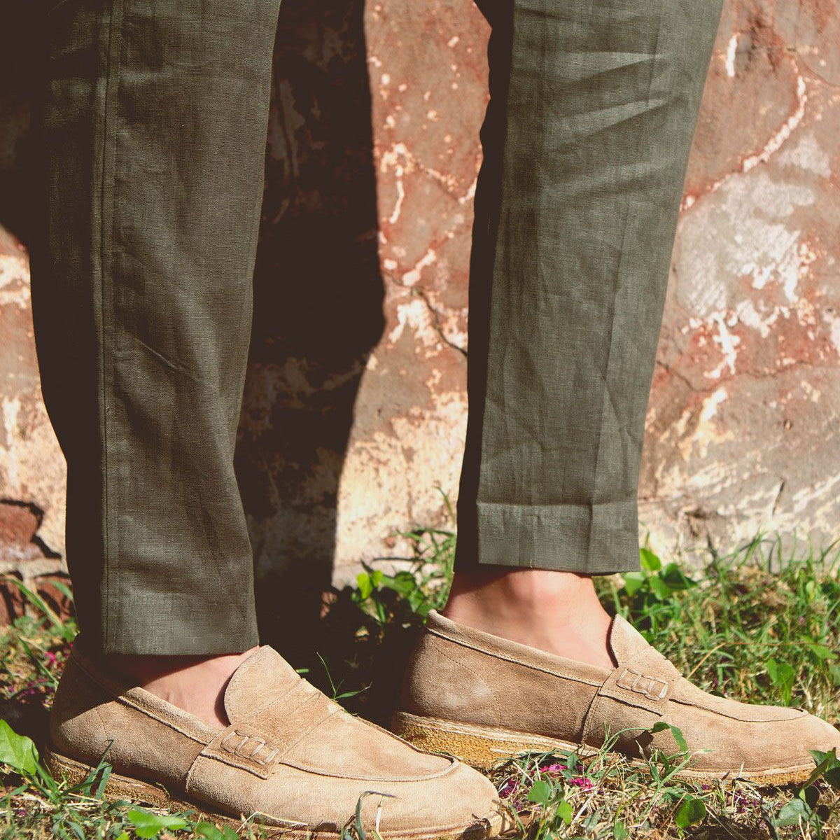 A person wearing green pants and tan suede Bed Stu Envoy loafers stands on grass with a rustic wall in the background, exuding Italian elegance.