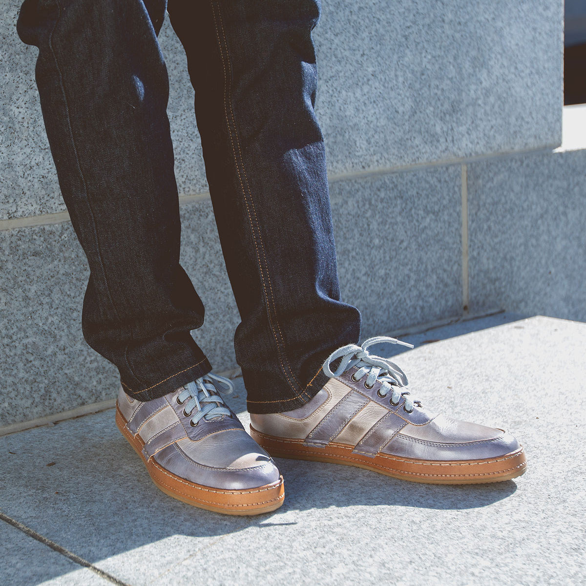 A person exudes casual cool in jeans and Bed Stu's Firepower gray men's sneakers, standing confidently on a stone surface, showcasing a classic vulcanized style.