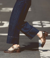 A person sporting flared blue denim pants and a pair of Forefront leather Mary Jane shoes by Bed Stu, known for their comfortable heel, walks on a street with large white painted markings.