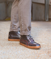 A person wearing Bed Stu's Formation brown high-top sneakers featuring subtle leather details, accompanied by beige pants, stands on a textured surface.