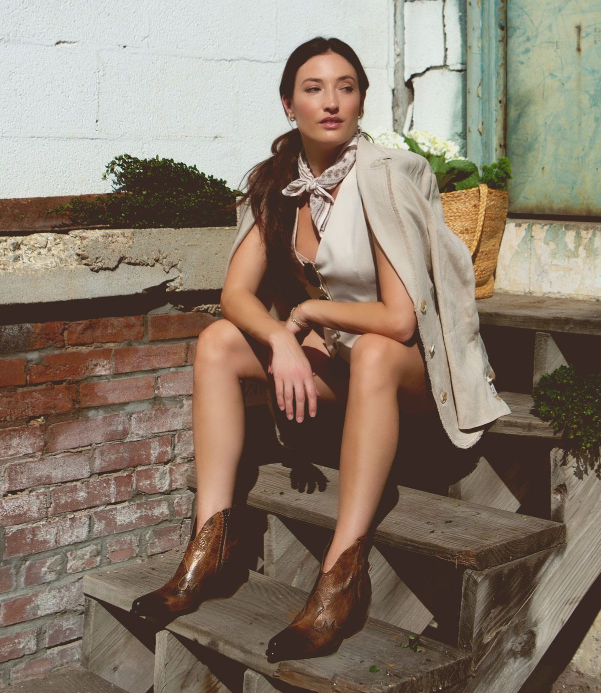 A person sits on outdoor wooden steps, dressed in a beige outfit with a scarf and brown boots. A blazer is draped over their shoulders. Plants and a woven bag are in the background, while their Furio Italian leather ankle booties from Bed Stu add a touch of handcrafted elegance from Italy.