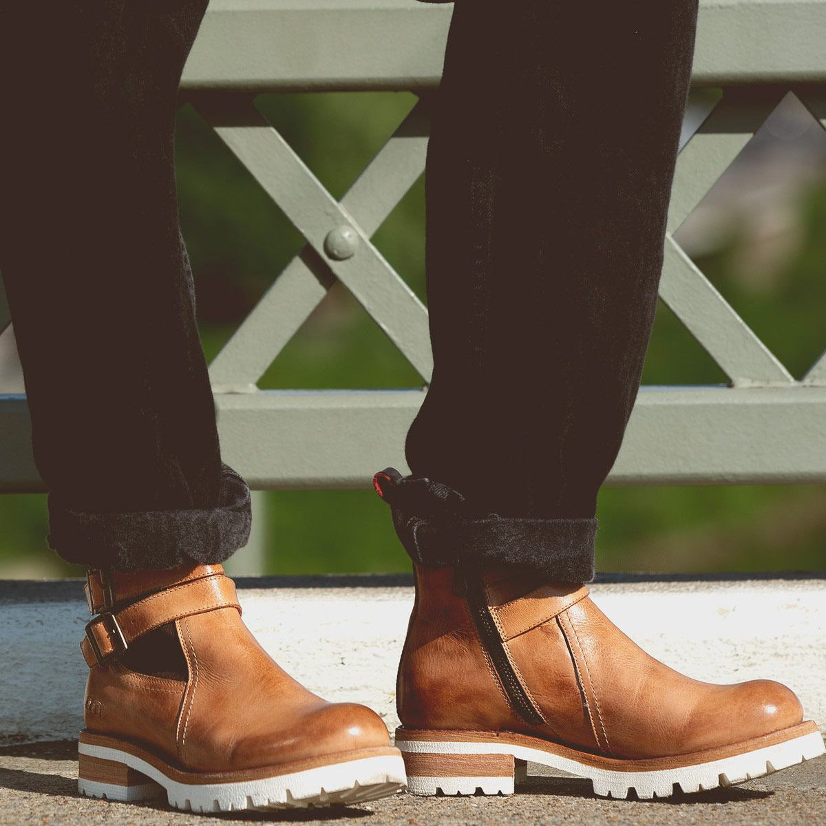 A knee-down shot of a woman in black cuffed denim standing on a brdige with bedStu Ginger II boots in tan on. 