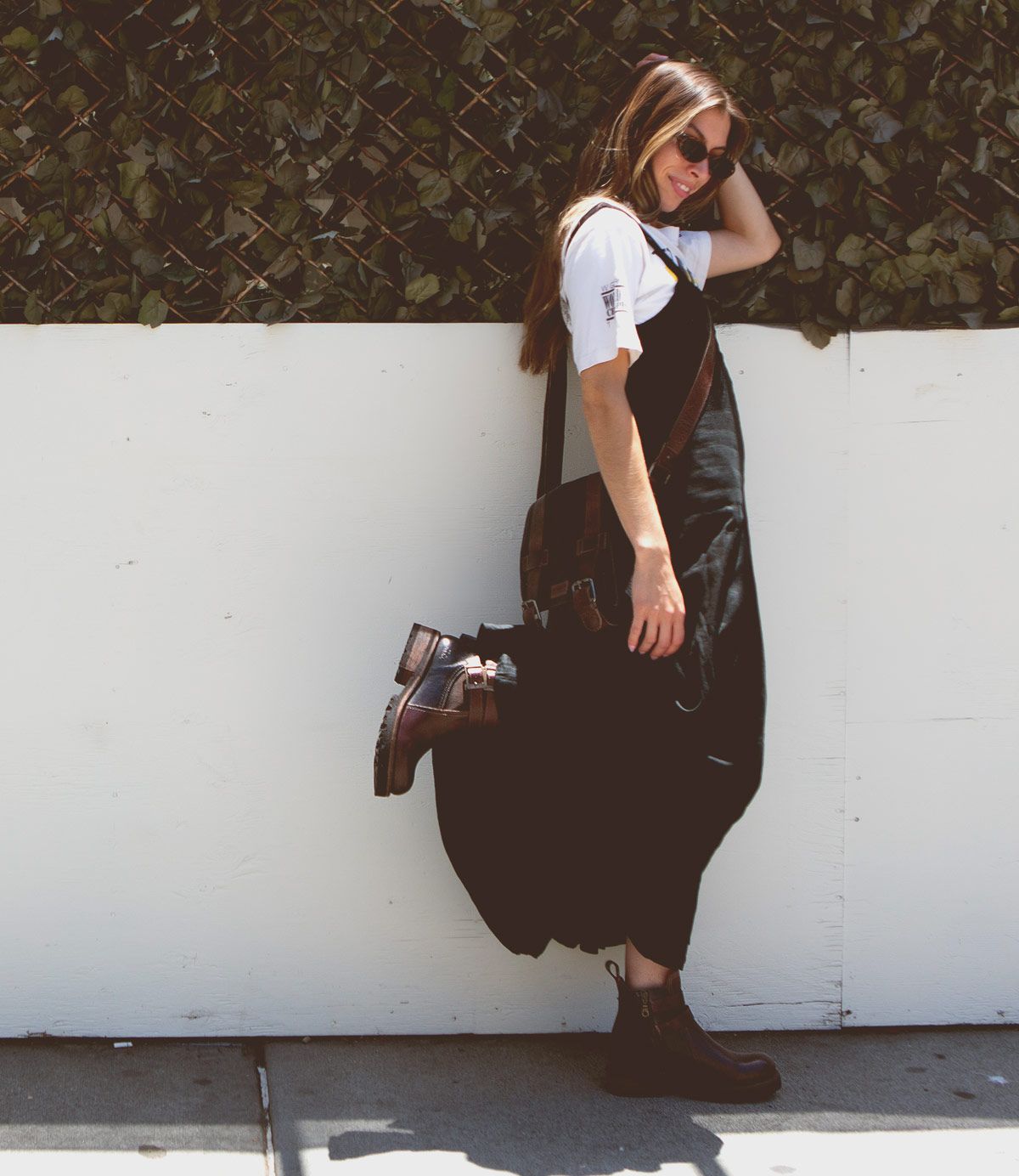 A person with long hair, sunglasses, and a white shirt under black overalls leans back against a white wall and a backdrop of brown leaves. They’re holding a bag and showcasing stylish touches like the Ginger II Chelsea ankle boots in brown with an inner zipper by Bed Stu.