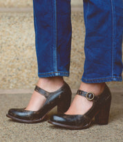 A person wearing dark brown Bed Stu Ishtar pumps with an adjustable strap and blue jeans stands on a concrete surface with stone steps in the background.