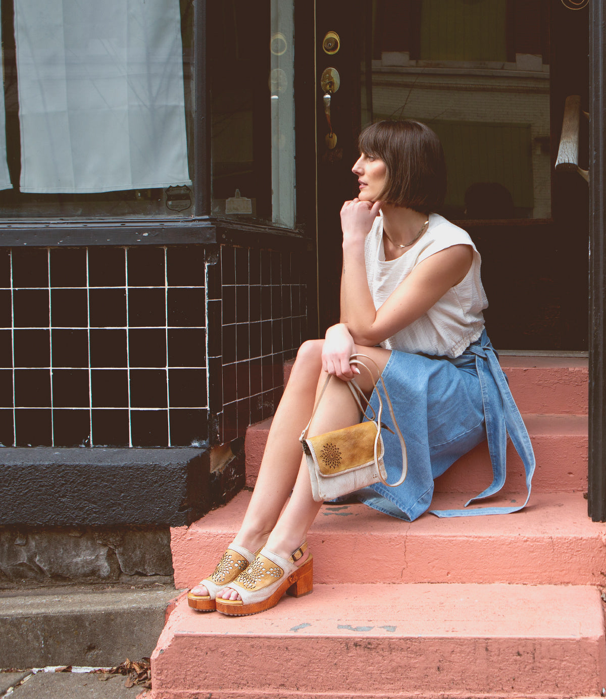 A woman sitting on the steps of a pink building, wearing Bed Stu Jinkie open-toe leather sandals with an adjustable ankle buckle.