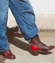 A person wearing blue jeans and brown Bed Stu Judgement boots with an 8-eye lace-up design is standing on a concrete surface in natural light.