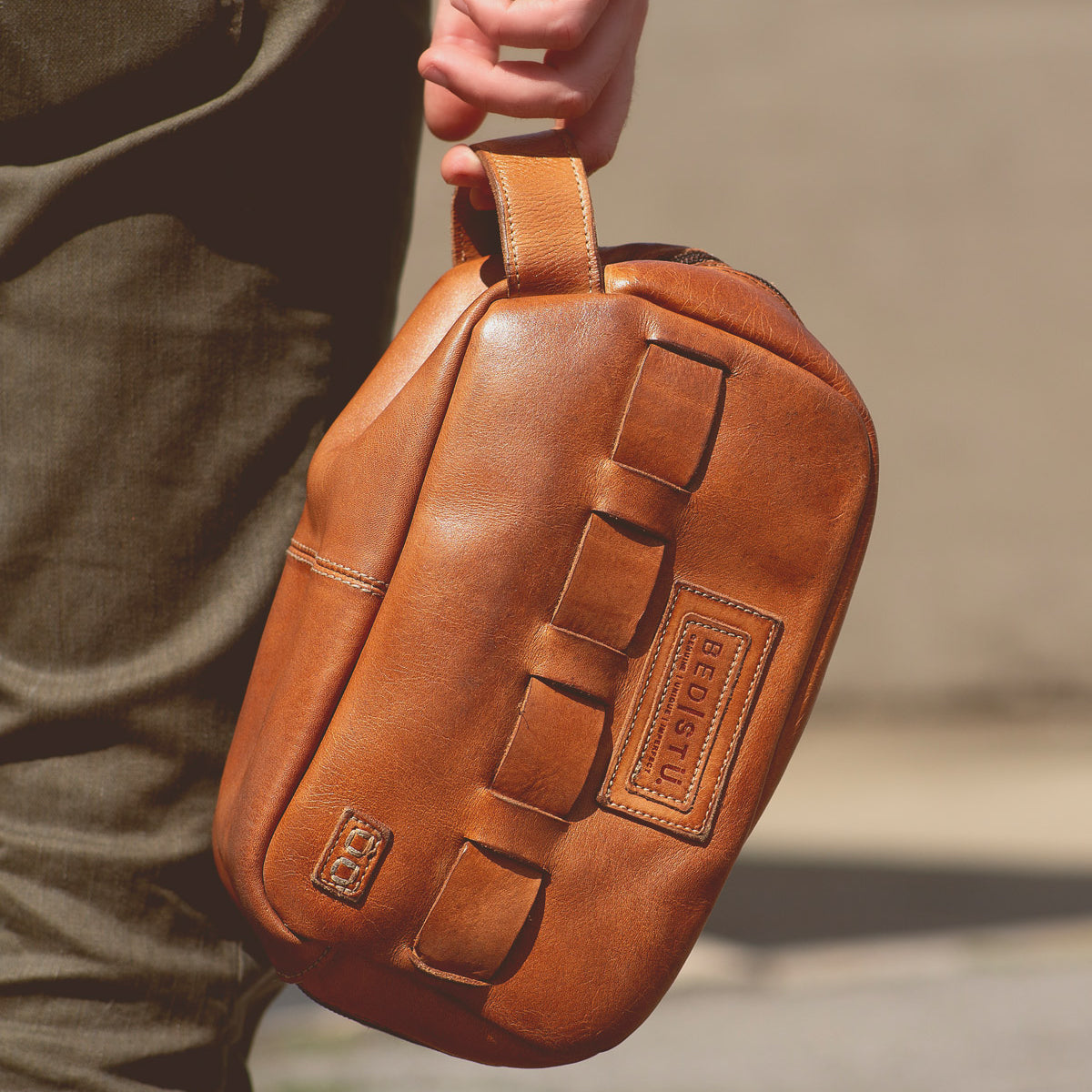 A person carrying a tan leather sling bag featuring a handle and multiple loops, with an embossed Bed Stu logo visible, perfect for travel essentials.