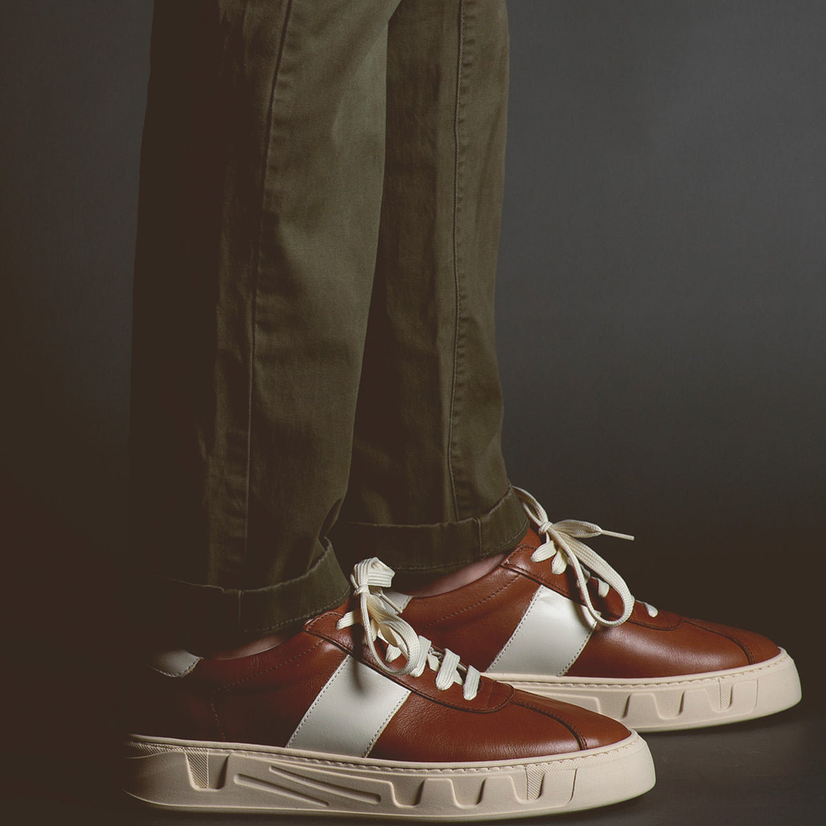 A person is wearing green pants and Bed Stu Law luxury leather sneakers with thick white soles, standing on a dark background.