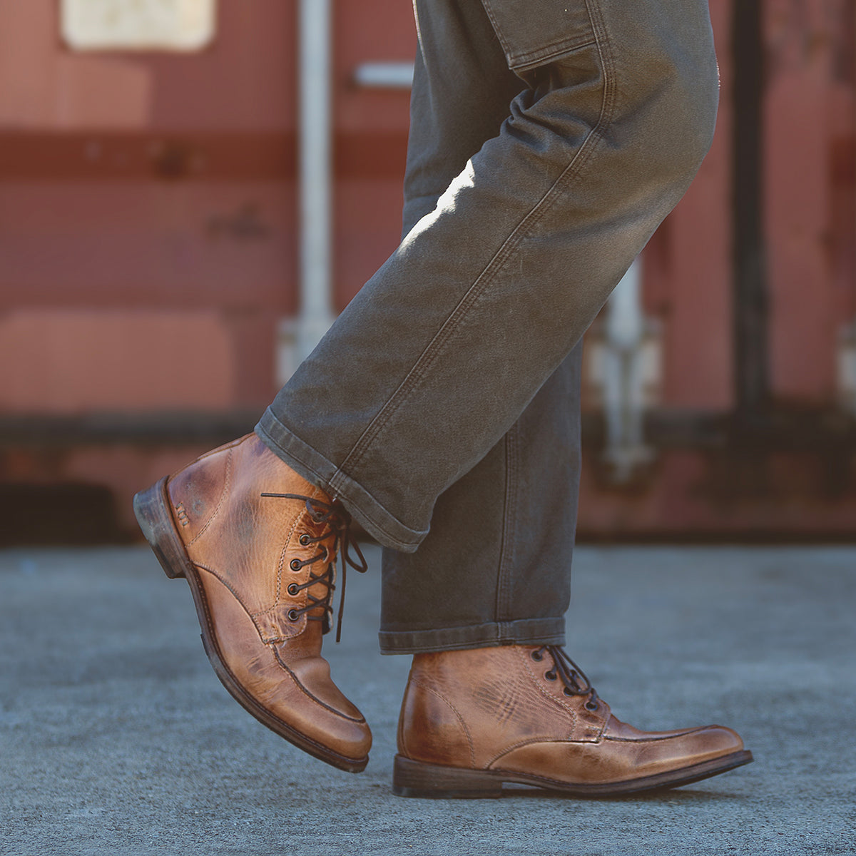 A person sporting a dressed-up look with the Lincoln II boots by Bed Stu and dark pants strides confidently on the concrete walkway.