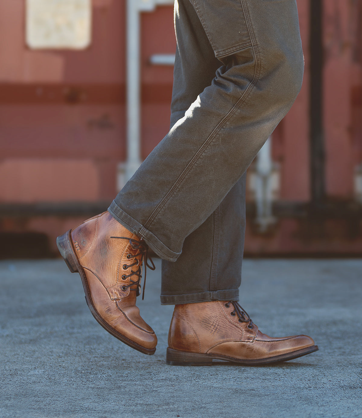 A person sporting a dressed-up look with the Lincoln II boots by Bed Stu and dark pants strides confidently on the concrete walkway.