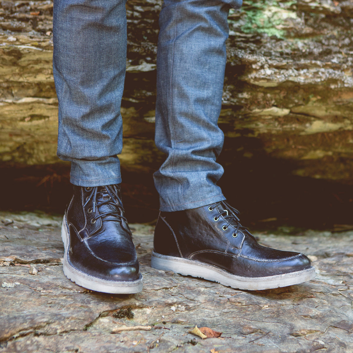 Close-up of a person wearing dark blue Lincoln jeans and Bed Stu black leather work boots standing on a rocky surface.