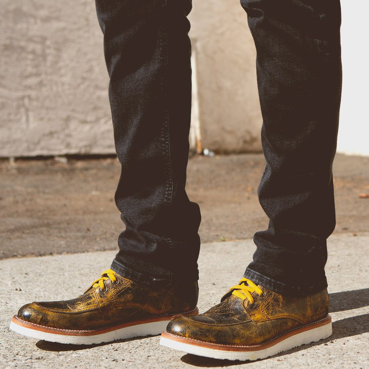 Person wearing black pants and metallic-colored Bed Stu Lincoln boots with yellow laces, standing outdoors on a concrete surface.