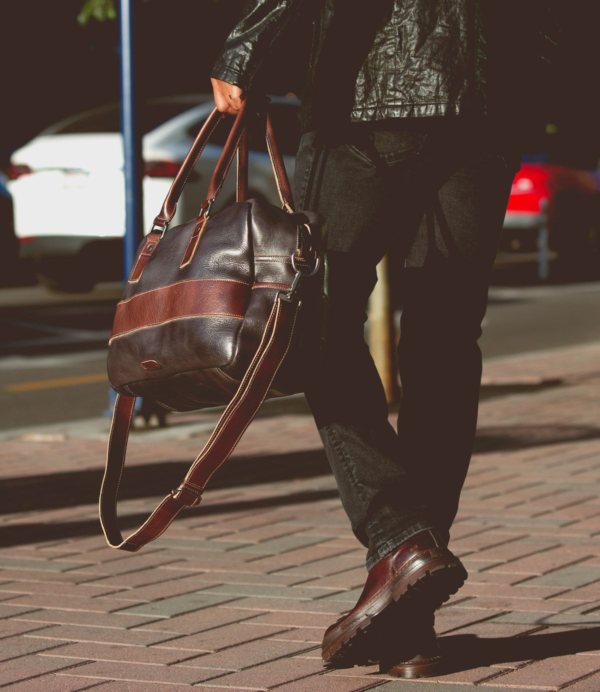 A person walking on a sidewalk is holding the Proem brown leather bag by Bed Stu, wearing a black leather jacket with a vintage metal belt buckle, black pants, and brown shoes.