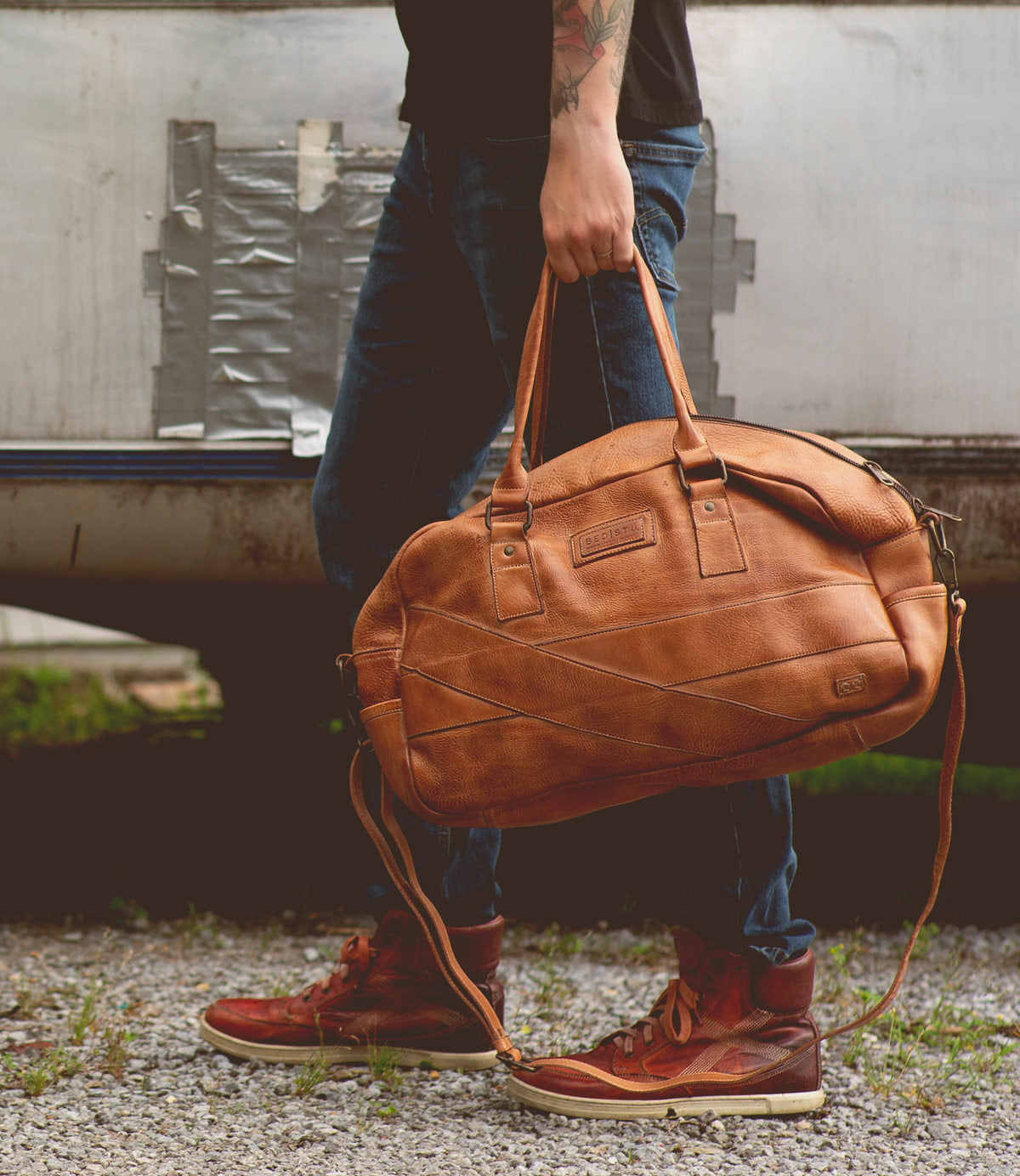 A person wearing jeans and red-brown sneakers holds a large brown leather duffel bag by the handles, standing outdoors on a gravel surface. This stylish travel companion, Luke by Bed Stu, features an adjustable crossbody strap for added convenience.