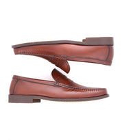 A pair of classic loafers crafted from high-quality leather, positioned horizontally with one on top and the other below, showcasing the side view and sole. These exquisite brown leather shoes, named Moat by Bed Stu, are handcrafted in Italy, epitomizing timeless elegance and superior craftsmanship.