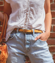 Person wearing a white t-shirt and blue jeans with a Bed Stu Monae II premium leather belt, holding a Bed Stu Monae II leather bag's strap.