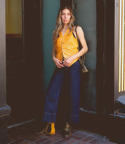 A person stands in a doorway wearing the Nandi yellow button-up vest from Bed Stu, dark wide-leg jeans, brown Chelsea boots, and a small shoulder bag.