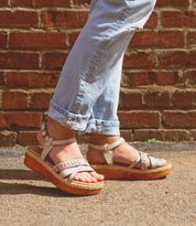 A person wearing Necessary light blue rolled-up jeans and Bed Stu strappy sandals with stitched straps walks by a brick wall.