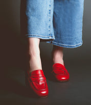 Person wearing blue jeans and red Bed Stu Paris penny loafers standing against a dark background.