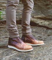 Person wearing Bed Stu Protege Light men's boots with cushioned leather insoles and beige pants, standing on a rocky surface outdoors.