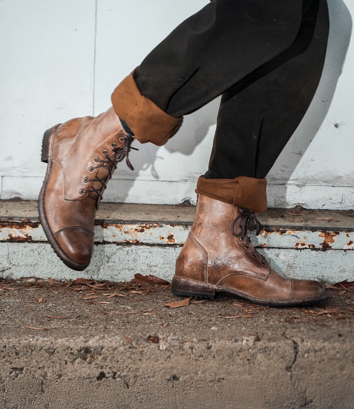 A person walks on a concrete surface near a metal wall wearing the Protege combat boots by Bed Stu, featuring a back zipper in brown, along with black pants.