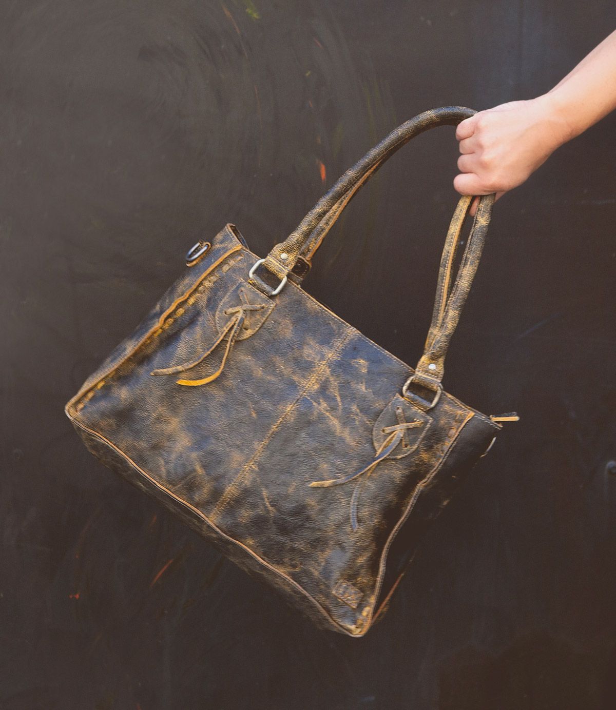 A hand holding the Bed Stu Rockababy distressed brown leather tote bag with two handles and decorative tassels against a dark background, perfect for an everyday bag.