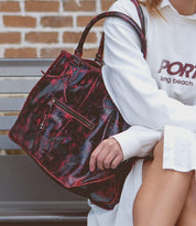 Person sitting on a bench holding a best-selling Rockaway handbag in a black and red pattern with a small front zipper compartment, wearing a white long-sleeve shirt. A brick wall is visible in the background, exemplifying the perfect blend of style and functionality characteristic of Bed Stu designs.