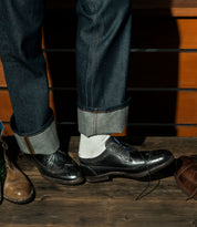 A person wearing cuffed jeans and black brogue shoes with white socks stands on a wooden floor, showcasing the intricate brogue details. Nearby, the Sandro II, a pair of handcrafted leather men's office shoes with a wingtip toe from Bed Stu, rests invitingly.