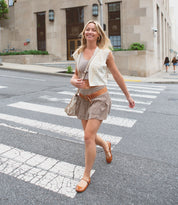 A woman in a beige outfit and brown shoes is walking across a city street crosswalk, smiling. She carries a small shoulder bag with an adjustable strap, the "Shuffle III" by Bed Stu, completing her chic look.