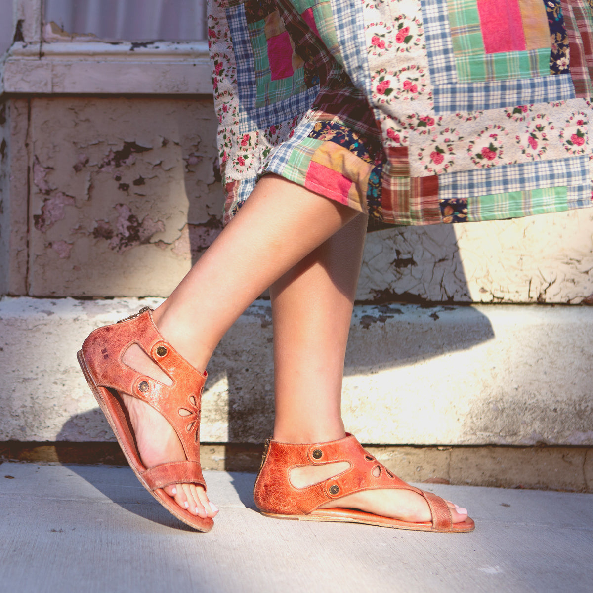 Woman wearing Bed Stu Soto Cutout flat leather sandals and patterned dress standing against a weathered wall.