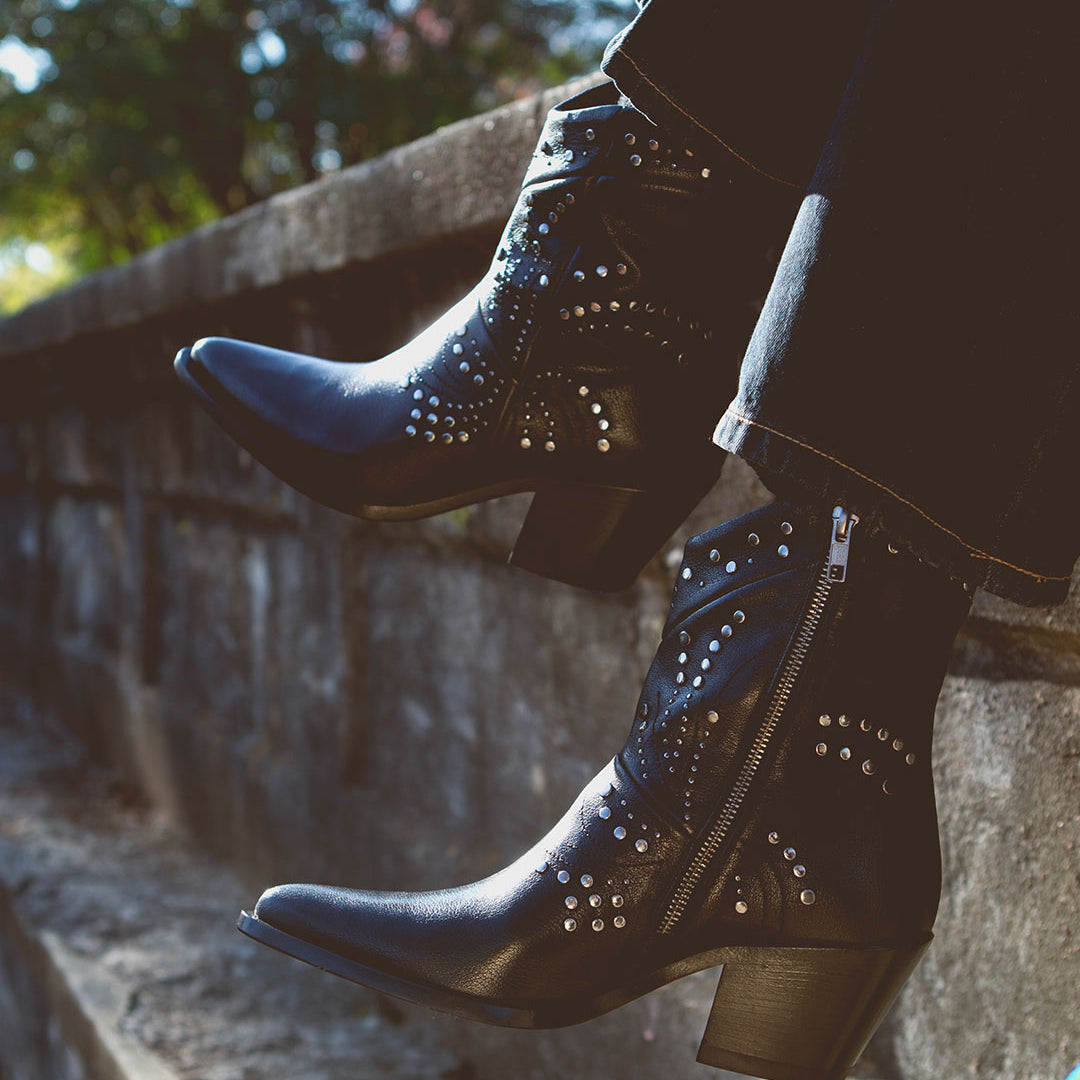 A person wearing Sposa booties by Bed Stu, crafted from black Italian leather with studded details and pointed toes, sits on a stone ledge.