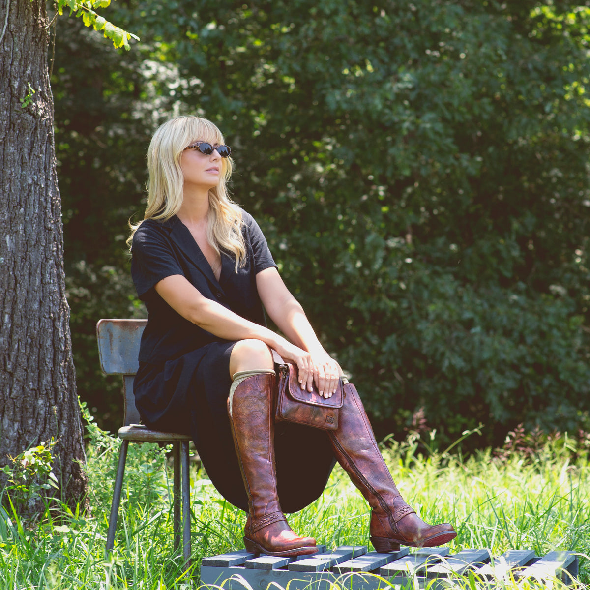 A woman in Takoma by Bed Stu, a western-shaped heel, is sitting on a bench in a field with leather inlays.