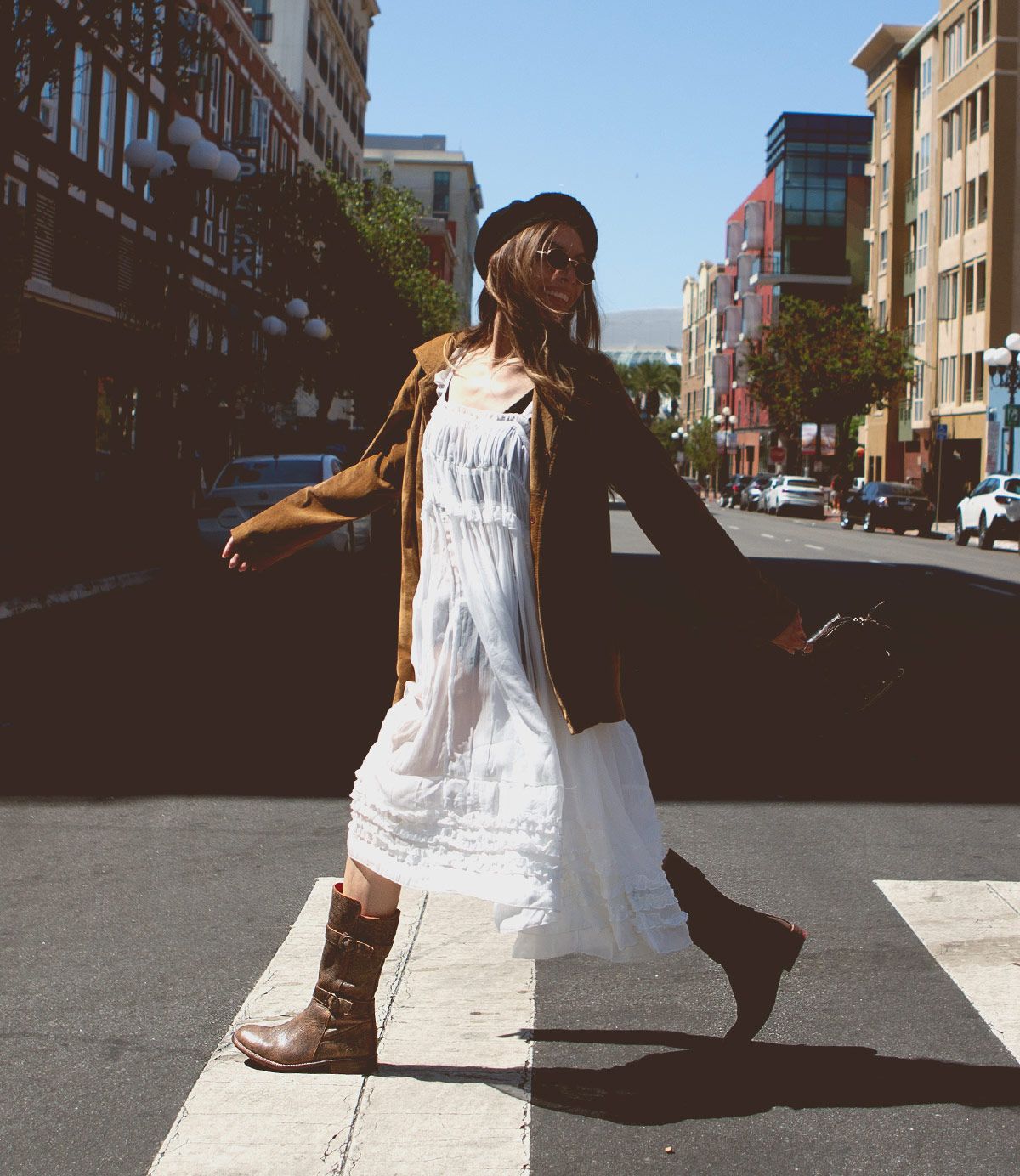 A person in a white dress, brown jacket, and Turn boots by Bed Stu crafted from vegetable-tanned leather walks across a city street on a sunny day.