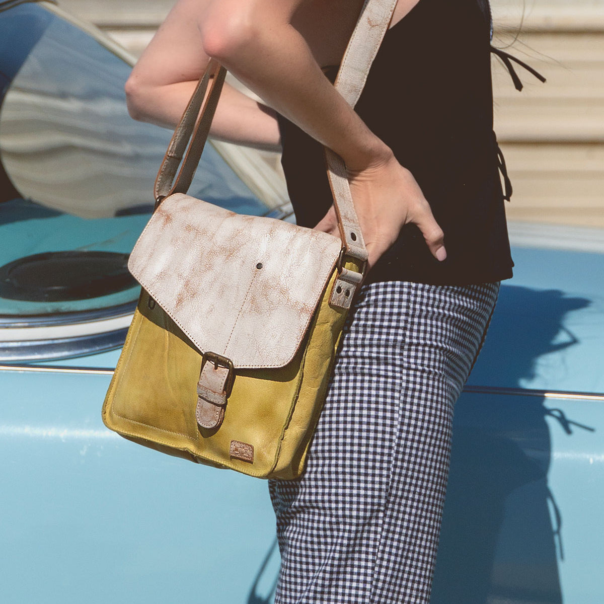 A person leans against a blue car, wearing a black top and checkered pants, holding the Venice Beach handmade leather crossbody bag by Bed Stu, featuring a bohemian-inspired design.