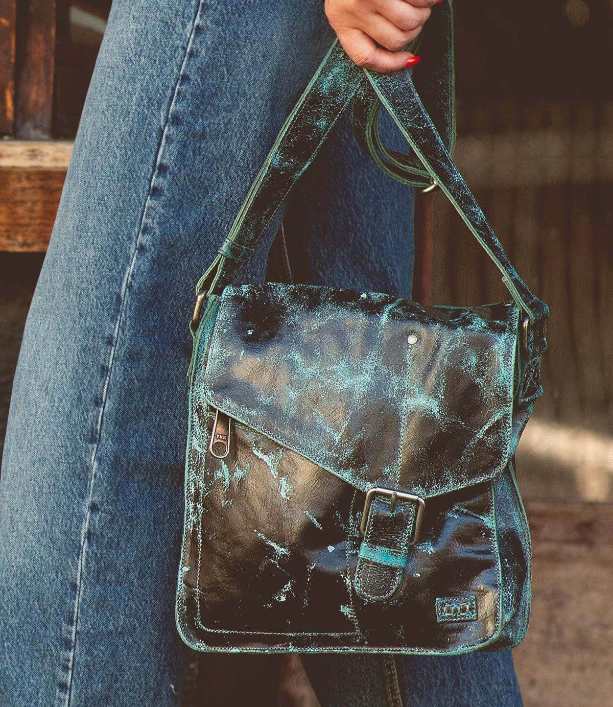 A person wearing blue jeans holds a Bed Stu Venice Beach handmade leather crossbody bag with an aged, distressed green and black leather finish and a buckle on the front flap.