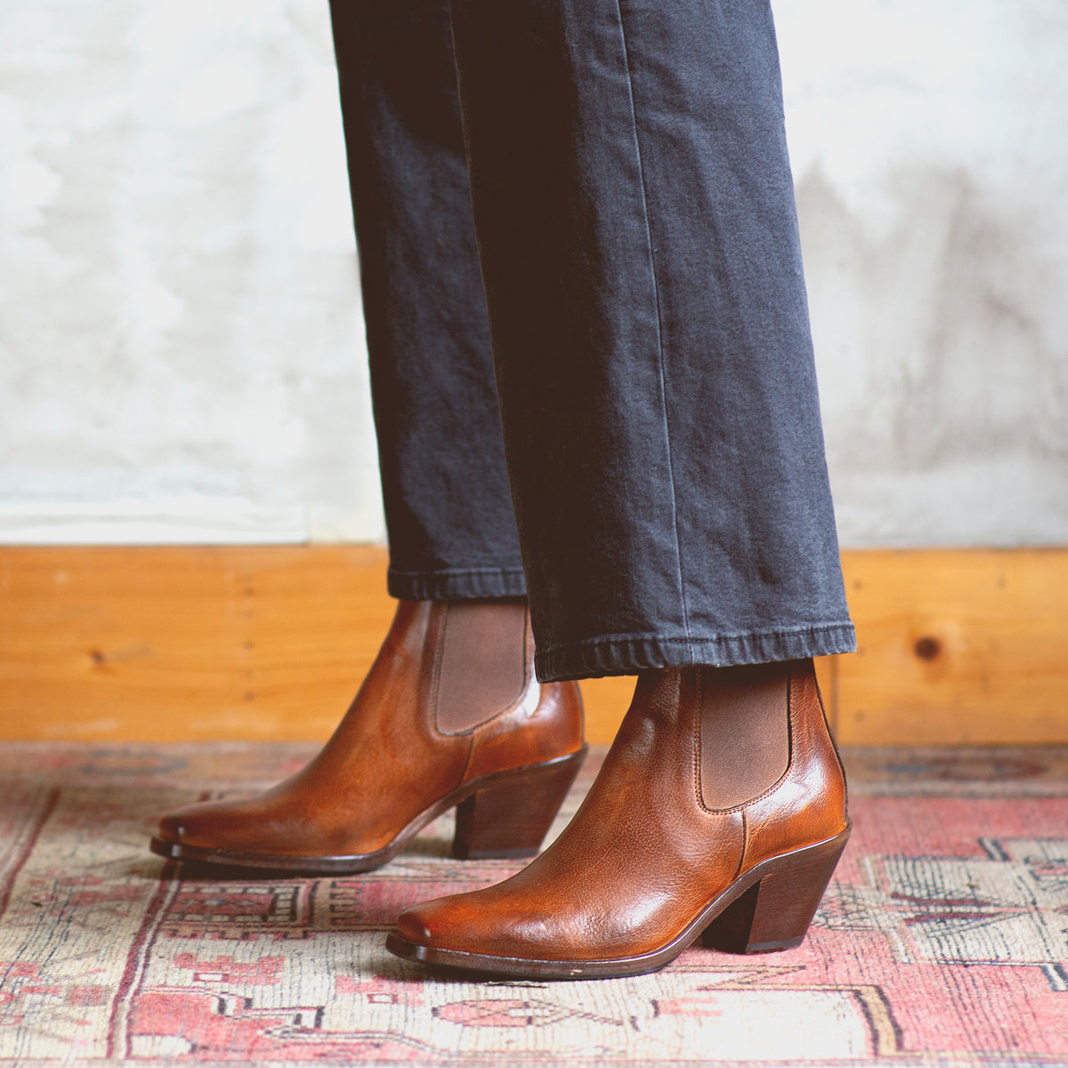 A woman exuding sartorial elegance wearing a pair of Bed Stu Viscera Chelsea boots on a plush rug.